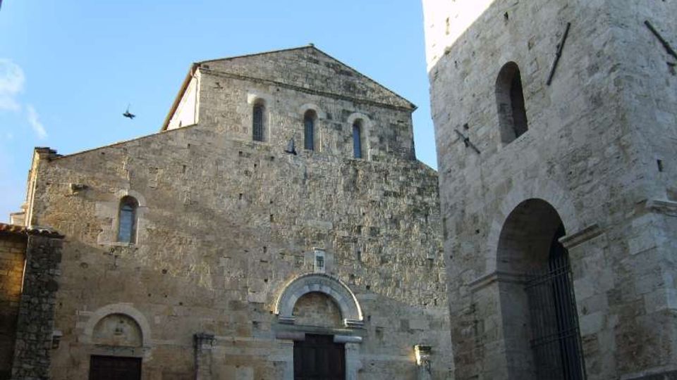 Facciata in pietra di una chiesa medievale con portali ad arco e piccole finestre, affacciata su una piazza. A destra si vede una torre campanaria in pietra con grande arco e apertura superiore, sullo sfondo di cielo sereno.
