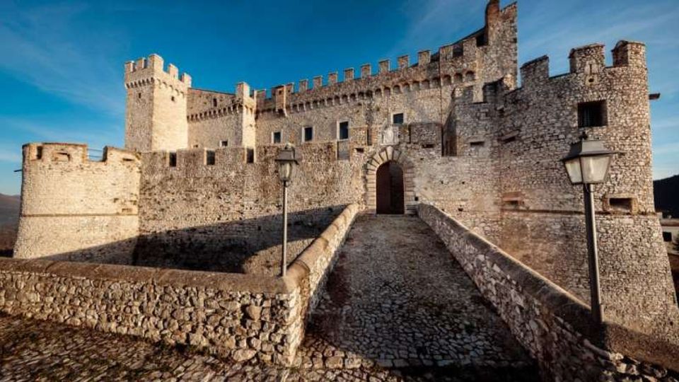 Castello medievale in pietra con mura merlate e torri, visto dall’esterno. Un ponte in pietra conduce all’ingresso ad arco, con lampioni lungo il percorso e cielo sereno sullo sfondo.