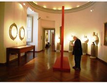 Interno di una galleria museale con pareti bianche e pavimento in parquet, illuminata da faretti a soffitto. Lungo le pareti sono esposti specchi con cornici dorate, una teca su tavolo e sculture su piedistalli, con un elemento espositivo rosso al centro della sala e un passaggio verso altre stanze sul fondo.