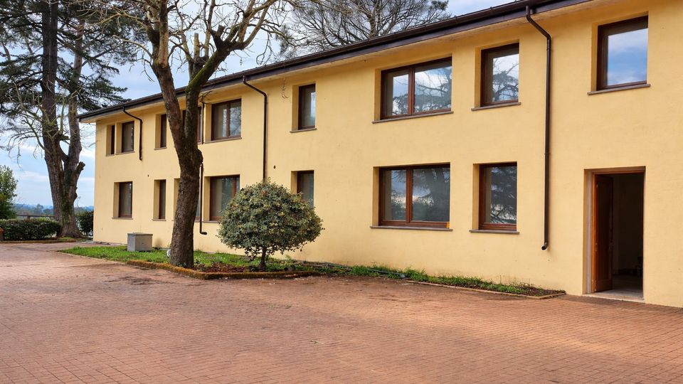 Edificio a due piani con facciata gialla e finestre rettangolari, affacciato su un ampio cortile pavimentato. Davanti alla struttura ci sono alberi e un cespuglio, con pluviali esterni e una porta d’ingresso aperta sul lato destro.