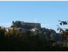 Panorama di un borgo su una collina, con un grande edificio storico fortificato in cima e file di cipressi lungo il crinale. In primo piano si vede un’area boschiva, con case in pietra sparse sui pendii sotto il castello.