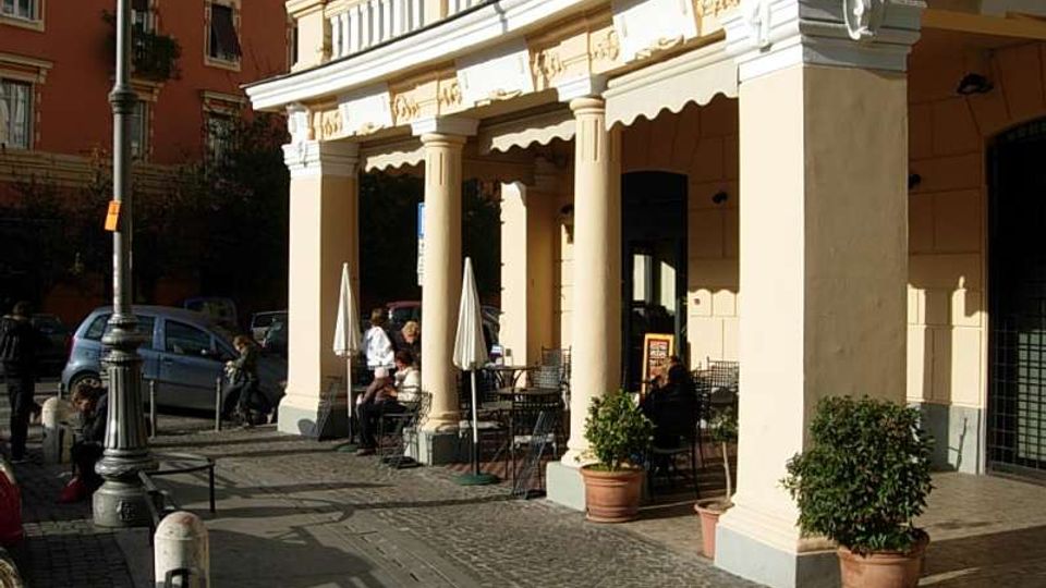 Portico curvo di un edificio in stile classico con grandi colonne chiare e decorazioni a rilievo, affacciato su un marciapiede in pietra. Sotto il portico si trovano tavolini e sedie di un bar con ombrelloni chiusi e vasi con piante, accanto a una strada con auto parcheggiate e lampione.