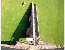 Veduta dall’alto di una lunga struttura in cemento a forma di rampa con gradini e pareti laterali, collocata al centro di un ampio prato verde. Alla base si vede una piattaforma con elementi metallici e un bordo in cemento che delimita l’area.