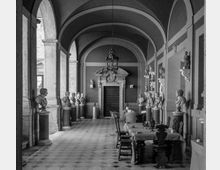 Corridoio interno con volte a botte e grandi archi in pietra, pavimento a piastrelle e una porta monumentale sul fondo. Lungo le pareti e su piedistalli sono esposti numerosi busti e statue, con tavoli e sedie disposti al centro sotto un lampadario.