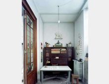 Piccolo studio interno con pareti bianche, grande armadio in legno scuro pieno di libri e oggetti e un tavolo bianco al centro illuminato da una lampada a sospensione. A sinistra c’è una porta in legno con vetri decorati, mentre a destra si vedono una parete con fogli appesi e un termosifone con telefono e sgabello.