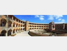 Cortile interno di un edificio storico a pianta semicircolare con due livelli di portici ad archi e pareti intonacate, affacciati su un’area centrale murata con vegetazione. Sul lato destro si vede una piccola struttura ottagonale con cupola e decorazioni, sotto un cielo azzurro con nuvole.