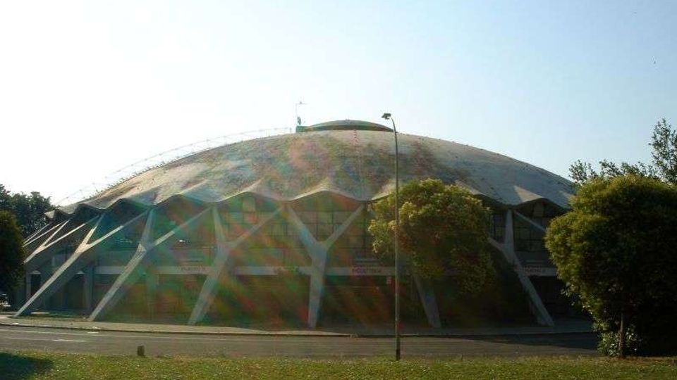 Grande edificio a cupola tipo palazzetto dello sport, con struttura esterna a travi triangolari e ampie vetrate alla base. Davanti si vedono una strada, un lampione e un’area verde con alberi ai lati.