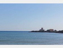 Veduta costiera con mare calmo in primo piano e, in lontananza, un piccolo promontorio con una torre in pietra e alcuni edifici. Cielo sereno sopra l’orizzonte marino.