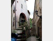 Vicolo stretto in un borgo con muri in pietra e intonaco, una scalinata che sale verso un grande arco d’ingresso e vasi di piante lungo i gradini. Sulla facciata opposta si vedono lampioni in ferro battuto e cavi sospesi, con vegetazione sullo sfondo tra gli edifici.