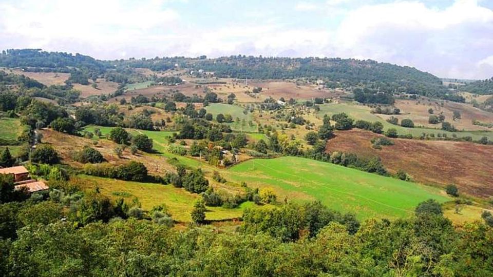 Panorama di campagna collinare con campi coltivati a mosaico e aree boschive, attraversate da strade e piccoli casali sparsi. In lontananza si vedono dorsali collinari sotto un cielo parzialmente nuvoloso.