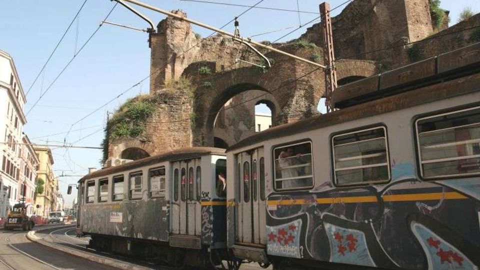 Tram urbano su binari in una strada cittadina, con cavi elettrici aerei e palazzi ai lati. Sullo sfondo si vedono grandi arcate e mura in mattoni di un antico edificio o viadotto, parzialmente coperto da vegetazione.