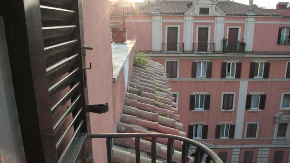 Vista da un piccolo balcone con ringhiera metallica e persiana in legno, affacciato su tetti in tegole e su un edificio urbano rosa a più piani con finestre e persiane scure. Sullo sfondo si vedono altri tetti e una luce di controluce.