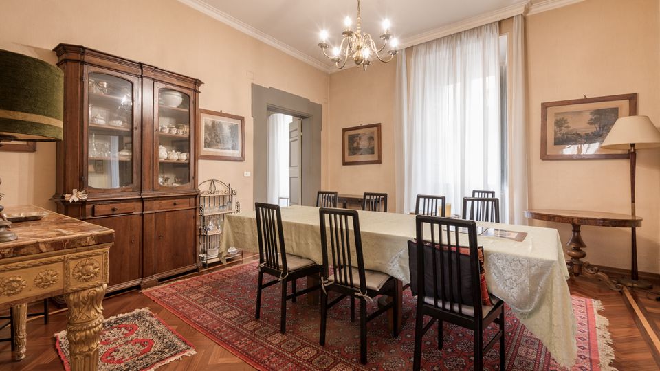 Sala da pranzo classica con grande tavolo rettangolare coperto da tovaglia e sei sedie, su tappeto persiano e pavimento in parquet. Lungo le pareti ci sono una credenza in legno con vetrina, quadri e una lampada da terra, con lampadario a soffitto e alta finestra con tende chiare.