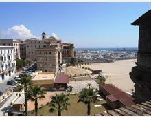Veduta panoramica di un porto turistico sul mare con numerose barche ormeggiate, affiancato da una grande piazza aperta e da edifici storici lungo il lungomare. In primo piano si vedono palme, piccoli edifici con tettoie e una strada costeggiata da palazzi.