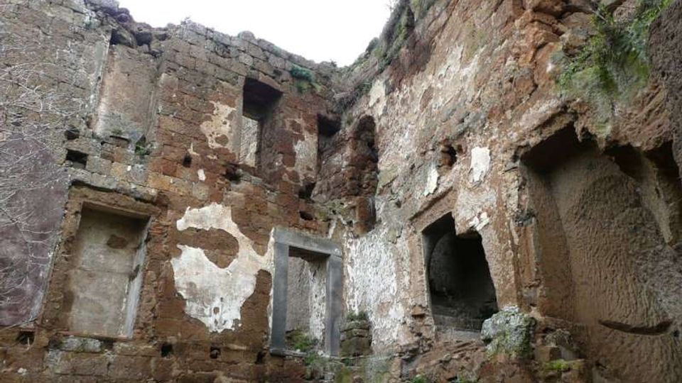 Ruderi di una struttura antica in pietra e tufo, con pareti parzialmente intonacate e aperture rettangolari e ad arco scavate nei muri. L’ambiente è a cielo aperto, con segni di erosione e vegetazione che cresce tra le pietre.