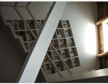 Interno di una stanza con una grande libreria a griglia piena di libri, vista in diagonale dietro una struttura metallica di scale o soppalco. A destra, una finestra con cornice in legno lascia entrare luce naturale sulla parete chiara.