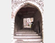 Passaggio ad arco in pietra che copre una scalinata lastricata in un vicolo stretto, con soffitto in legno e pareti in muratura grezza. In fondo si intravedono case in pietra con finestre e piante in vaso lungo i gradini.