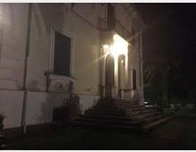 Facciata esterna di un edificio a più piani con intonaco chiaro, ingresso illuminato da una lampada e una scalinata in pietra che conduce a un portico con archi. Davanti si vede un piccolo giardino con cespugli e vegetazione, in un contesto notturno.