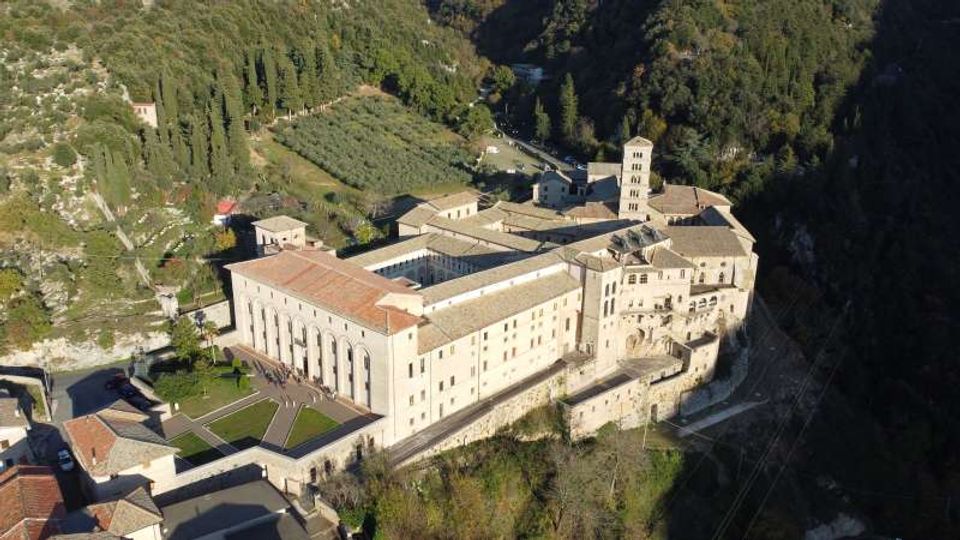 Veduta aerea di un grande complesso monastico in pietra con tetti in coppi e un campanile quadrato, disposto attorno a cortili e terrazze su un pendio. L’edificio è circondato da colline boscose e campi coltivati, con strade e piccoli edifici rurali nelle vicinanze.
