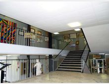 Atrio interno di un edificio pubblico con pavimento in marmo chiaro, ampio vano scala centrale con ringhiere metalliche e un pianerottolo su due livelli. Pareti decorate con pannelli colorati e cornici, illuminazione a plafoniere e porte vetrate sullo sfondo.