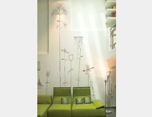 Soggiorno moderno a doppia altezza con parete bianca decorata da grandi disegni stilizzati di fiori verticali e divani modulari verde lime. A destra si vede un soppalco con scaffali e ringhiera, illuminato dalla luce naturale dall’alto, con lampade da terra in metallo.