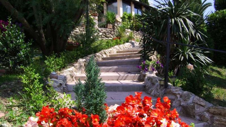 Sentiero con gradini in pietra che sale attraverso un giardino terrazzato ricco di vegetazione mediterranea, con aiuole di fiori rossi in primo piano. In cima si vede una casa con grandi finestre, circondata da muretti a secco e alberi.