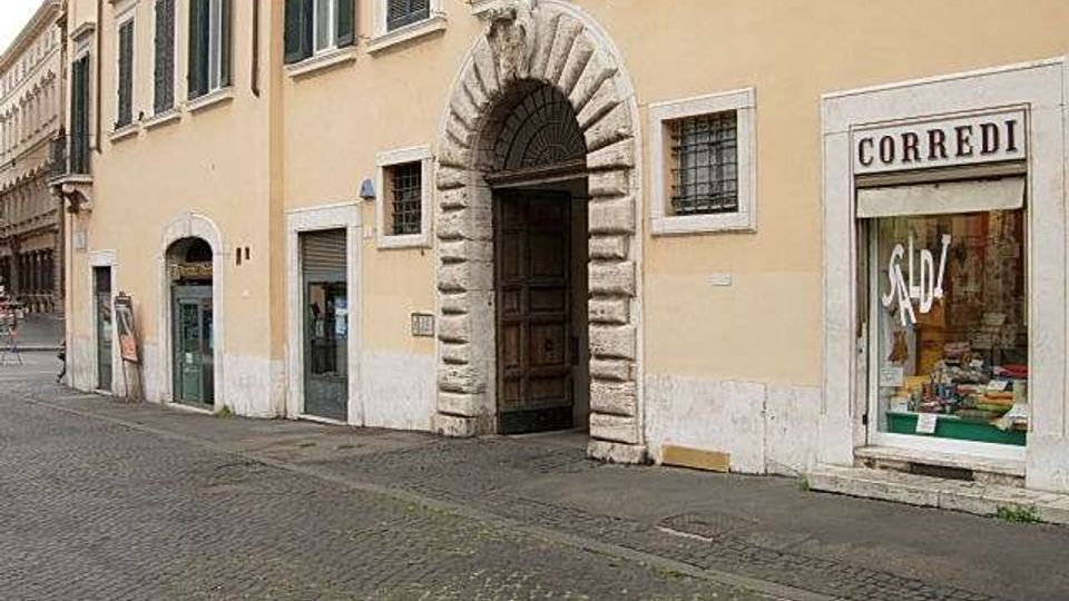 Facciata di un edificio storico color ocra lungo una strada lastricata, con grande portale ad arco in pietra bugnata e finestre con persiane. A destra si vede una vetrina di negozio con insegna “CORREDI” e scritta “SALDI”.