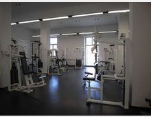 Ampia sala palestra interna con pavimento scuro in gomma, pareti bianche e file di macchine per pesi e cavi disposte lungo i lati. Il locale è illuminato da lunghe lampade fluorescenti a soffitto e da grandi finestre sul fondo.