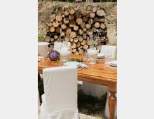 Tavolo da pranzo in legno apparecchiato all’aperto con piatti bianchi, calici, brocche e vasi in ceramica con fiori, circondato da sedie con coperture bianche. Sullo sfondo si vede una catasta di tronchi tagliati contro una scarpata di terra.