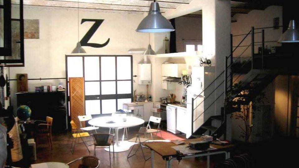 Interno di un loft open space con cucina a vista, tavoli e sedie in stile industriale, grandi lampade a sospensione e soffitto con travi a vista. Sul lato destro una scala metallica sale a un soppalco, con pareti bianche e un grande elemento grafico a forma di “Z” sulla parete.