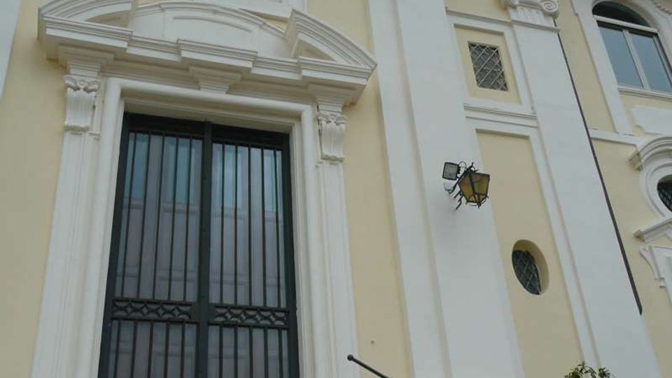 Facciata di un edificio storico color crema con cornici e lesene bianche decorate, grande portale con grata metallica e scalinata in pietra con corrimano. In alto si vede una finestra ad arco e una torre campanaria parzialmente visibile sul tetto.