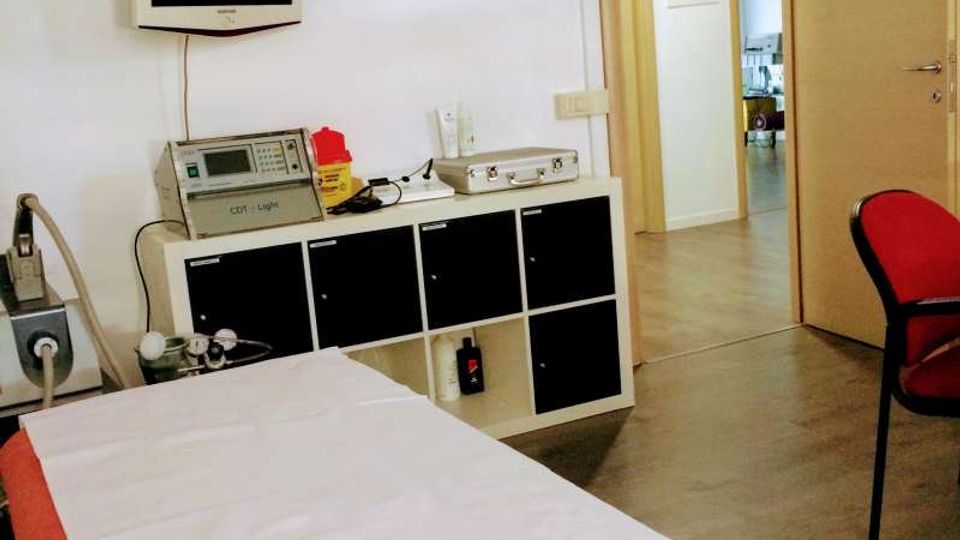 Ambulatorio medico con lettino da visita coperto da lenzuolo, lampada chirurgica a soffitto e monitor a parete sopra un mobile con attrezzature. Porta aperta verso un corridoio interno e pavimento in linoleum chiaro.