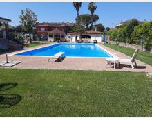 Piscina rettangolare all’aperto con bordo piastrellato e acqua azzurra, circondata da prato curato e lettini. Sullo sfondo si vedono un gazebo e una piccola struttura con portico ad archi e tetto in coppi, con palme e case residenziali intorno.