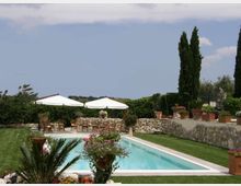 Piscina rettangolare all’aperto con bordo chiaro, circondata da prato curato e muretti in pietra con vasi di fiori. Sullo sfondo ci sono due grandi ombrelloni bianchi con sedie, siepi e alti cipressi sotto un cielo parzialmente nuvoloso.