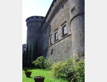 Vista dal basso di un castello in pietra con alta torre circolare e mura massicce, con finestre rettangolari e mensole sotto il tetto. In primo piano c’è un prato con cespugli, cipressi e alcuni grandi vasi in terracotta.