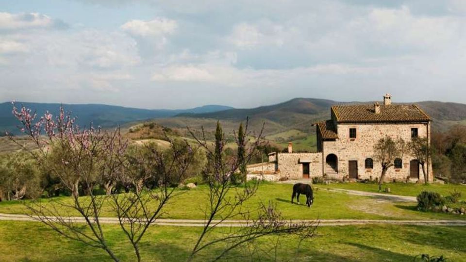 Casale rurale in pietra con tetto in tegole e archi al piano terra, affacciato su un ampio prato attraversato da un sentiero. Intorno ci sono alberi spogli o in fiore e colline ondulate sullo sfondo sotto un cielo parzialmente nuvoloso.