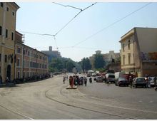 Ampia piazza urbana con binari del tram che curvano in primo piano e cavi elettrici sospesi sopra la strada. Ai lati si vedono edifici a più piani con facciate colorate e finestre, marciapiedi con veicoli parcheggiati e, sullo sfondo, una collina con un edificio storico.