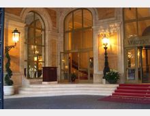 Ingresso di un hotel con facciata in pietra, grandi finestre e porte ad arco in vetro e gradini in marmo. Due lampioni a parete illuminano il portico, con tappeto rosso sulla scalinata e vasi con piante ai lati.