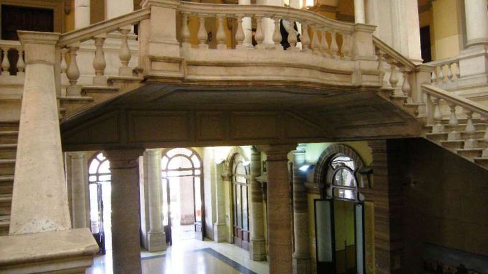 Interno di un edificio storico con grande scala in pietra e ballatoio con balaustra, sostenuti da colonne. Al piano terra si vedono archi e porte vetrate ad arco che si affacciano su un atrio luminoso.