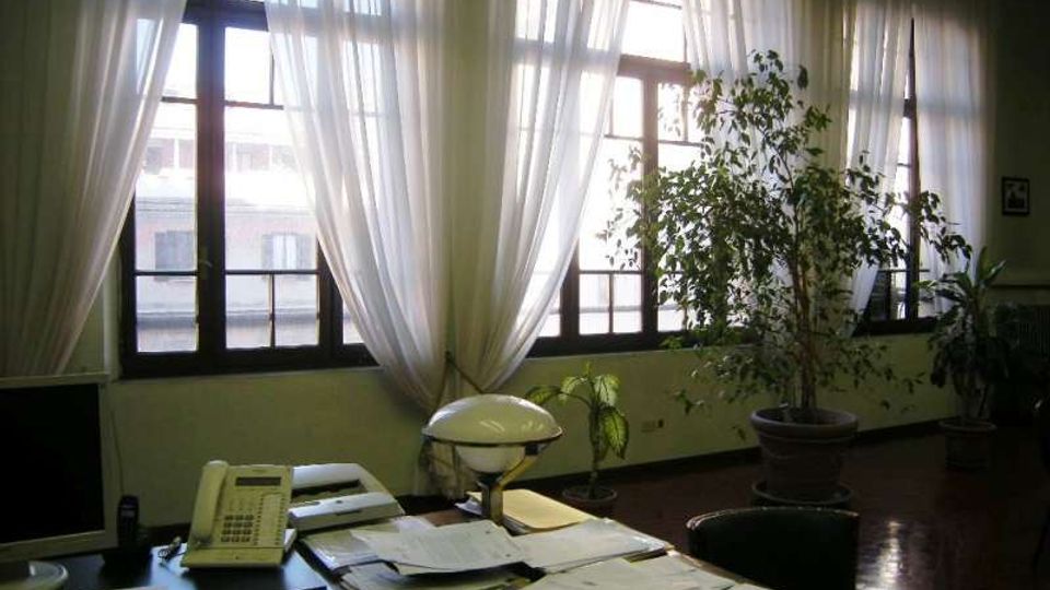 Ufficio interno con grande scrivania in primo piano coperta di documenti, telefono e lampada da tavolo. Sullo sfondo ampie finestre con tende bianche trasparenti e diverse piante in vaso lungo la parete.