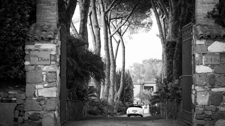 Ingresso di una villa con due pilastri in pietra e cancelli in ferro, che si apre su un vialetto acciottolato fiancheggiato da vegetazione e alti alberi. In fondo si intravede l’edificio della villa e un’auto parcheggiata lungo il viale.
