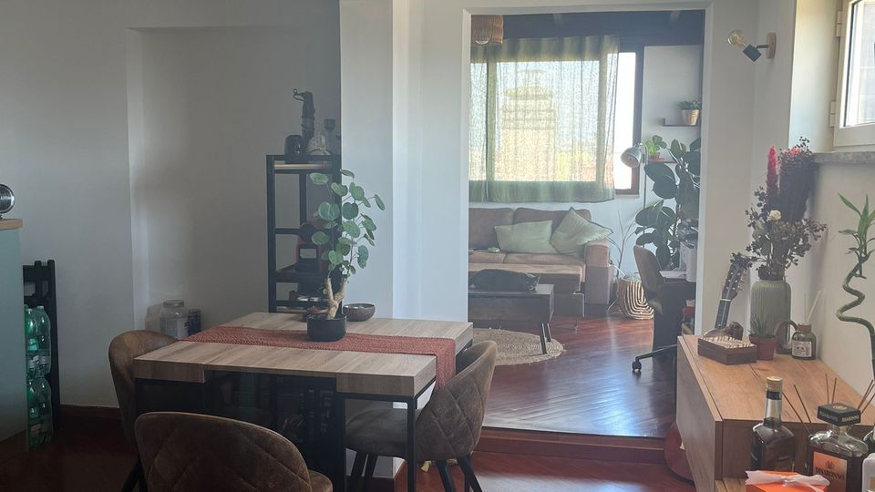 Interno di un appartamento con zona pranzo open space, tavolo rettangolare con quattro sedie su pavimento in legno scuro e pareti bianche. Sullo sfondo, un piccolo salotto rialzato con divano e grande finestra con tende, affiancato da piante e mobili bassi lungo la parete.