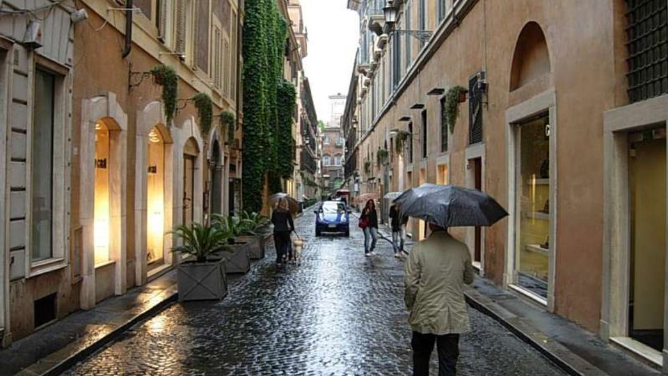 Strada stretta di centro storico con pavimentazione in sampietrini bagnati dalla pioggia, fiancheggiata da edifici alti con vetrine, finestre con persiane e alcuni balconi. Lungo il percorso si vedono lampioni a parete, grandi fioriere e una facciata coperta di edera; sullo sfondo la via prosegue tra le case.