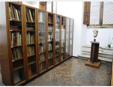 Stanza interna tipo biblioteca o archivio con pavimento in piastrelle e pareti bianche, fiancheggiata da alte librerie in legno con ante in vetro piene di volumi. In fondo si vedono una finestra con inferriate, alcuni gradini in pietra e un busto su piedistallo.