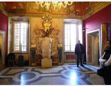 Interno di una sala storica decorata con affreschi e stucchi, con due grandi finestre e un lampadario a soffitto. Al centro della parete principale si trova una nicchia con scultura e busti laterali, mentre il pavimento presenta motivi geometrici in pietra.