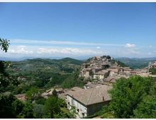 Veduta panoramica di un borgo collinare con case in pietra dai tetti in coppi, disposto lungo un crinale. Intorno si estendono colline verdi e campi coltivati sotto un cielo azzurro con nuvole all’orizzonte.