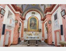 Interno di una chiesa con navata centrale che conduce a un altare in marmo bianco, sovrastato da una grande pala dipinta entro un’arca dorata e un lampadario. Le pareti laterali hanno colonne e cornici in marmo rosato con balconate bianche, mentre il soffitto è a volta decorata con motivi dipinti.