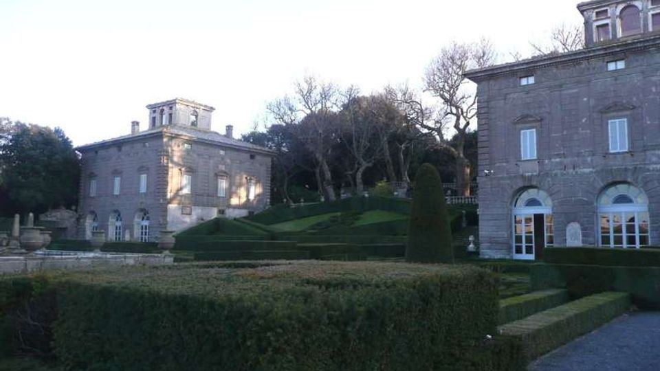 Giardino formale con siepi geometriche e vialetto in ghiaia davanti a edifici in pietra grigia in stile villa storica, con finestre ad arco e porte vetrate. Sullo sfondo si vedono prati terrazzati e alberi spogli lungo un pendio.