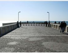 Passeggiata su un molo o pontile lastricato in pietra, con parapetti laterali e due lampioni, che termina con una ringhiera davanti al mare. Sullo sfondo si vede l’orizzonte marino sotto un cielo limpido.