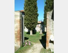 Ingresso di un piccolo cimitero tra pilastri in mattoni e cemento, con un vialetto su prato che conduce a un cipresso alto e a loculi sullo sfondo. Ai lati si vedono altri cipressi e un’aiuola di fiori rosa vicino al passaggio.